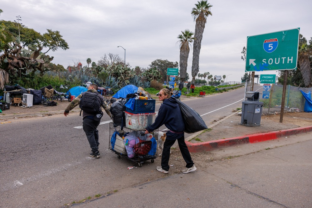 Two people push a cart filled with items across freeway toward encampment
