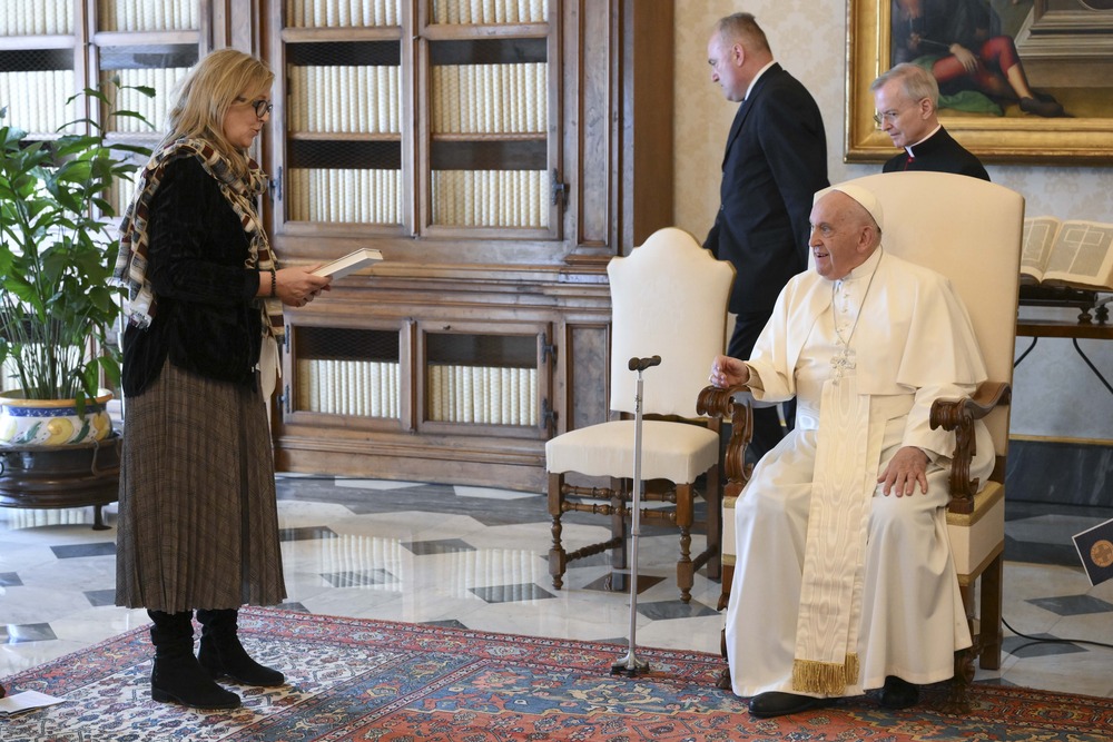 Pope Francis, seated, smiles while listening to Maeve Heaney speak.