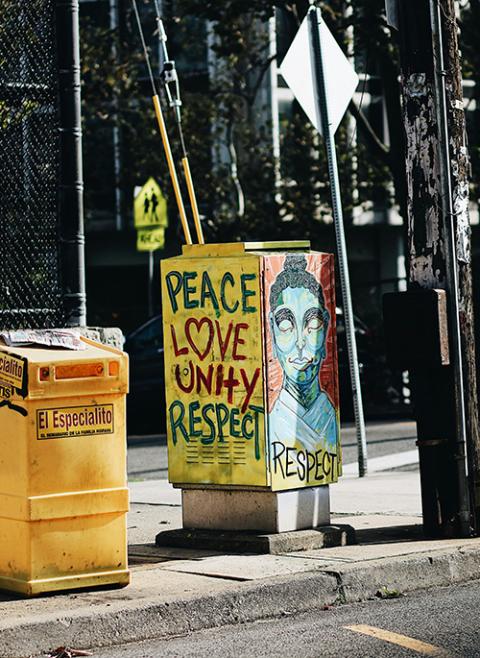 An outdoor photo displays a yellow trash bin that is covered with graffiti that includes the words "peace," "love," "unity" and "respect." (Unsplash/Domo .)