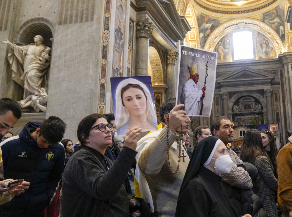 A man holds a rosary and a picture of Benedict that says "Te Esperabamos" in the middle of a crowded line