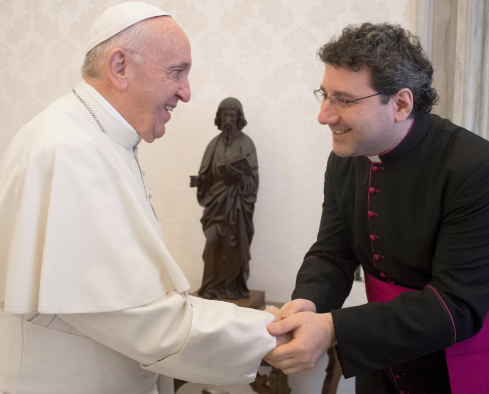 Pope Francis shakes a man's hand who is wearing glasses and a black and purple cassock