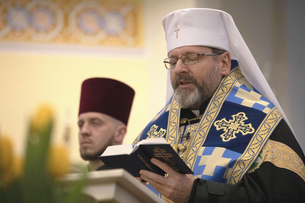 An older white man with glasses wears a round white hat with a cross and a train and a blue and gold stole with crosses on it. He reads from a book.