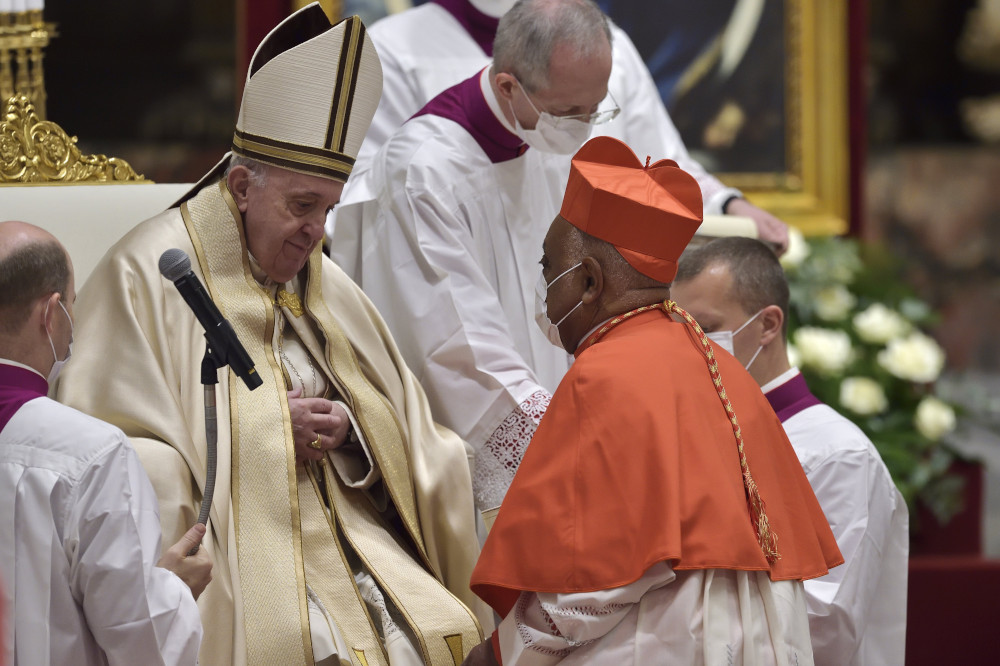 Cardinal Gregory, wearing a mask and a red biretta, kneels in front of Pope Francis, wearing a mitre