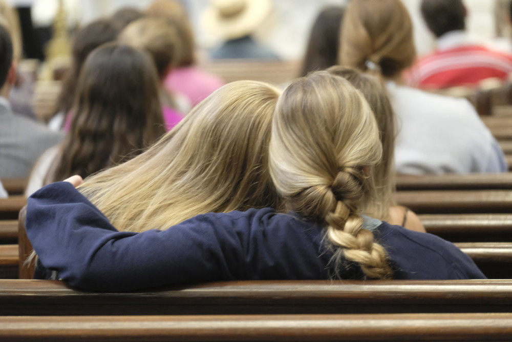 A person with long blond hair leans on a person with a blond braid, who wraps her arm around the other person. They sit in a church pew.