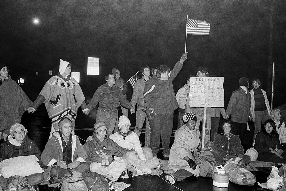 Antinuclear protesters sit on the ground in an attempt to block a gate at Seneca Army Depot in Romulus, New York, as counter demonstrators join hands and wave a flag behind them on Oct. 25, 1983. (AP/Ron Frehm)