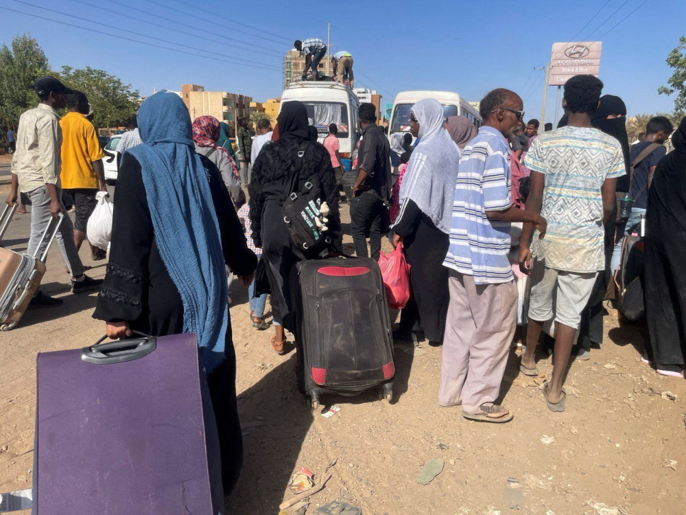 A group of people with suitcases gather outside white buses. Some women wear headscarves