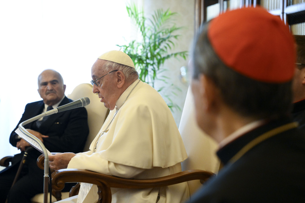 Pope Francis speaks into a microphone with a man in a suit and a man in a red zucchetto looking on