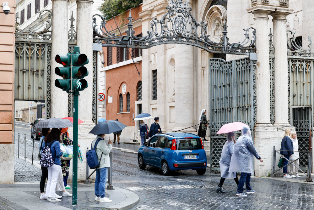 A small blue car drives through an ornate wrought iron gate. People walk by with umbrellas and rain ponchos.