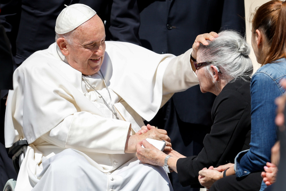 Pope Francis, sitting in his wheelchair, places a hand on the head and hands of an older white woman with a hearing aid