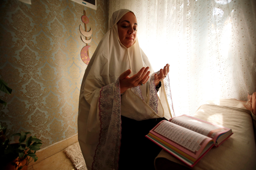 A woman wearing a waist-length hijab lifts her hands, palms raised, while sitting in front of a book with Arabic writing