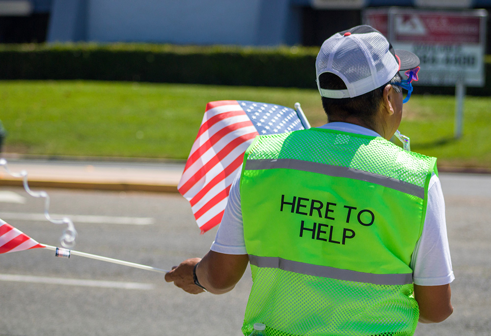The back of a person on a street, holding American flags and wearing a green vest that says, "HERE TO HELP." (Unsplash/Elissa Garcia)