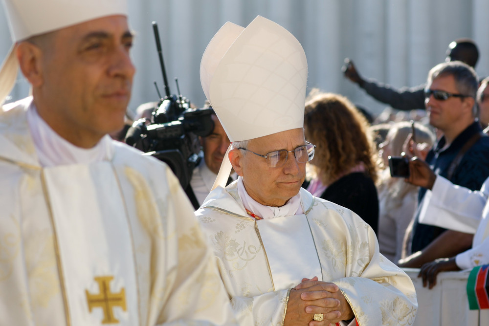 Two light-skinned men wear white mitres and vestments as they stand side-by-side with people and cameras in the background
