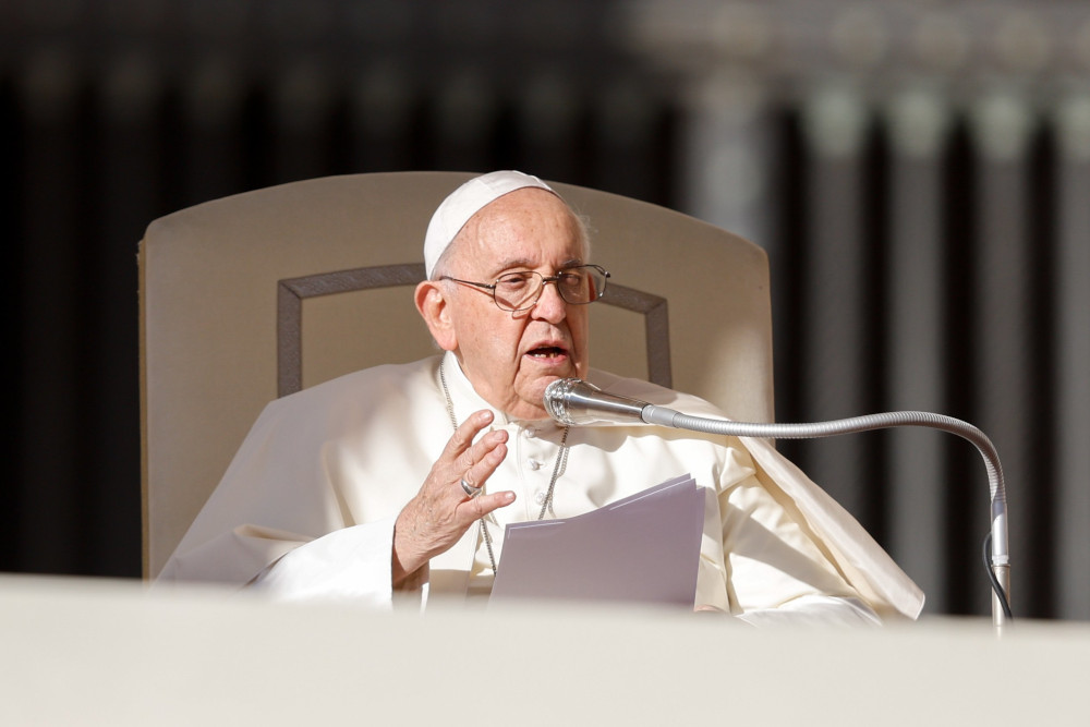 Pope Francis speaks into a microphone while sitting in a white chair and holding a paper