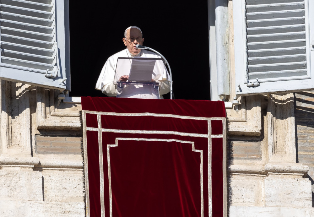 Pope Francis reads from a sheet of paper while standing in shadow in the window of his apartment
