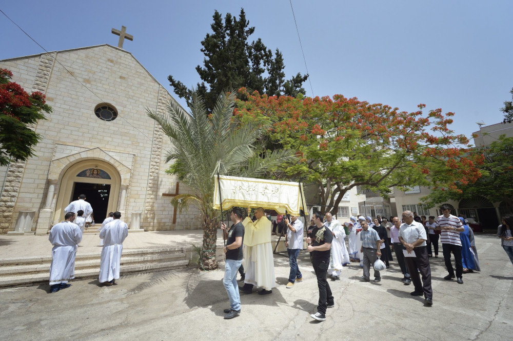 A group of people walk into a stone church. In the middle, four people hold a canopy that covers a priest holding a monstrance.