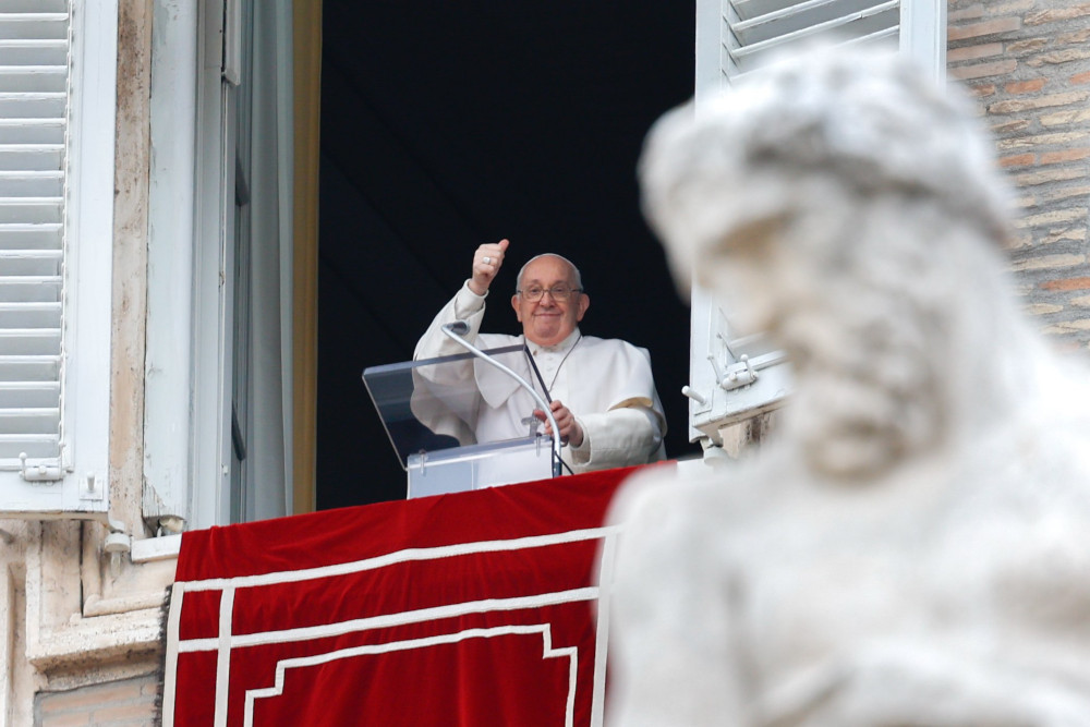 Pope Francis raises a thumbs up from his apartment window. A statue is visible in front of the shot of the window.