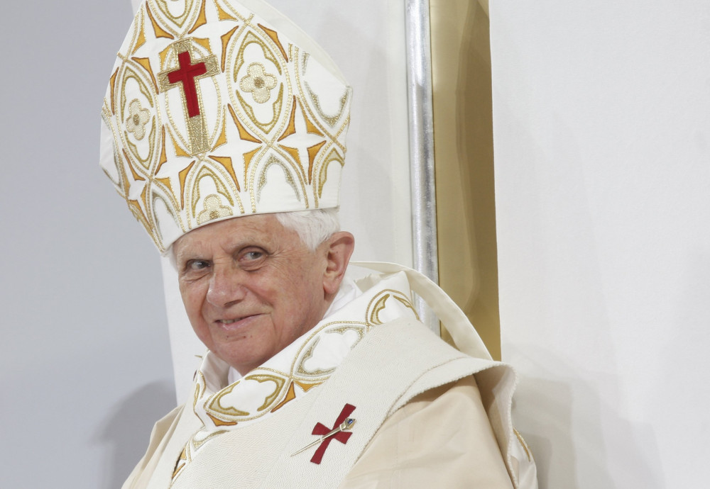 Pope Benedict XVI wears a white and gold mitre with a red cross on it and matching vestments