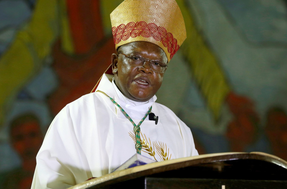 A Black man wearing glasses, a gold and red mitre and white vestments speaks behind a podium