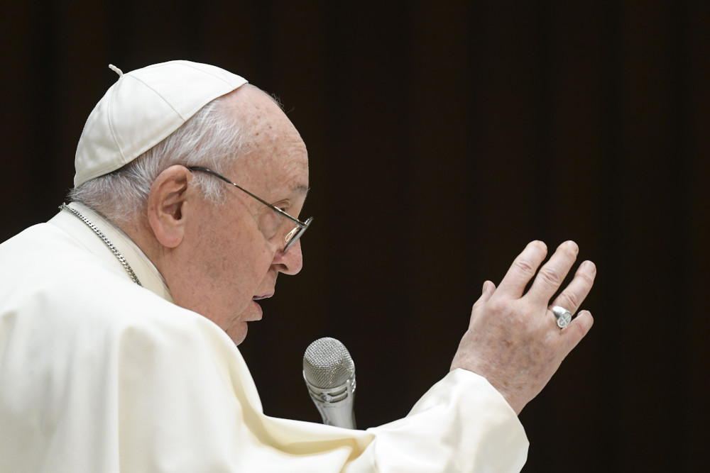 Pope Francis raises his right hand as he speaks into a microphone. He is against a black background.