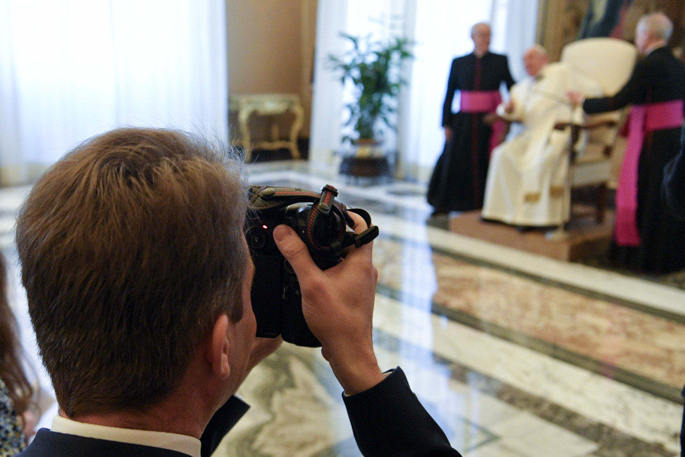 A white man raises a camera. Pope Francis and two bishops can be seen blurry in the background.