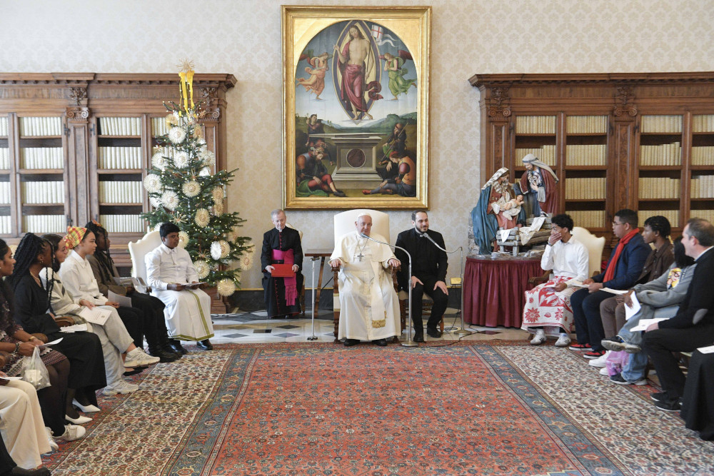 Pope Francis sits in a chair in a carpeted room with bookshelves. Attendees, including many young people of color, sit on the sides of the room.