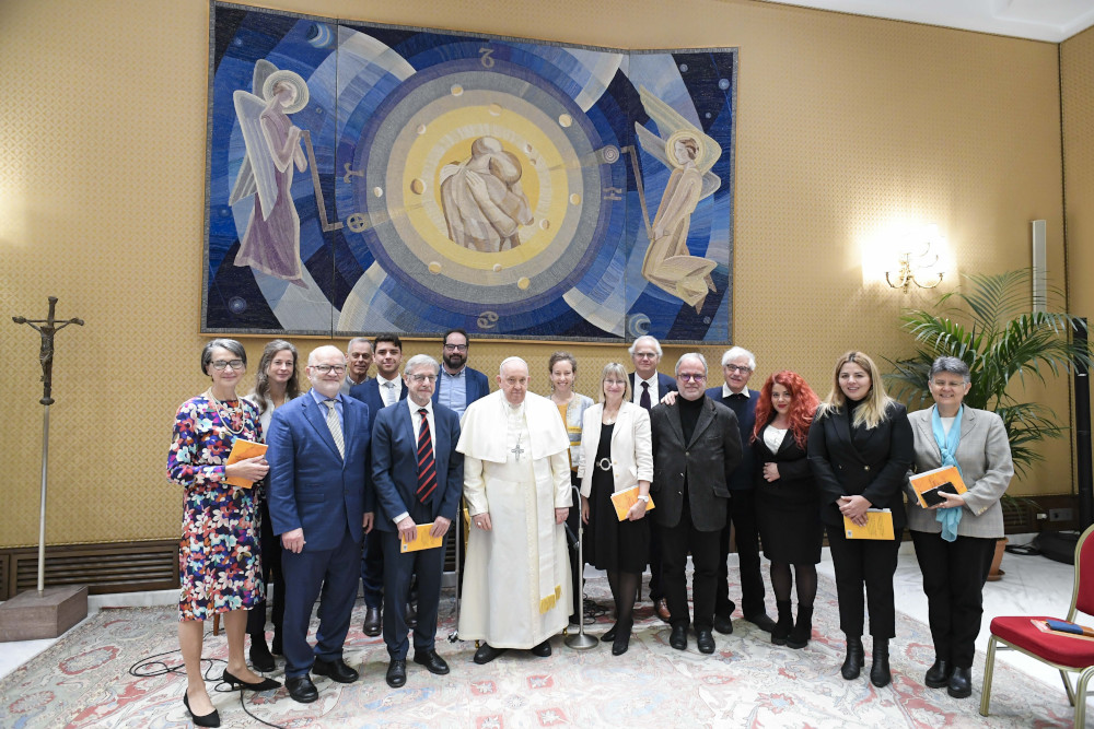 Pope Francis and a group of people pose for a photo below an abstract tapestry