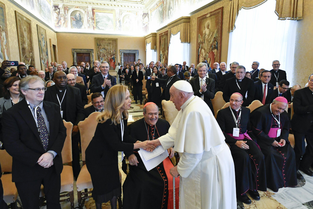 Pope Francis shakes hands with a blond woman in business clothes in front of a hall full of people