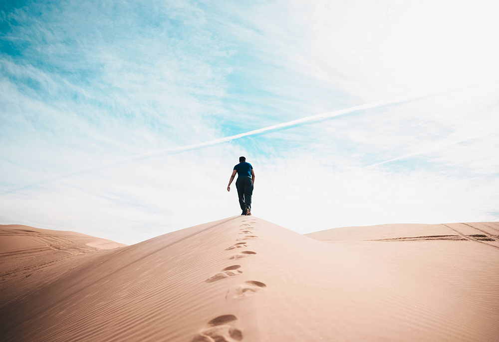 A person walks up a hill of sand in a desert setting, with a blue sky in the background, and footsteps behind the person walking.(Unsplash/Nathan McBride)