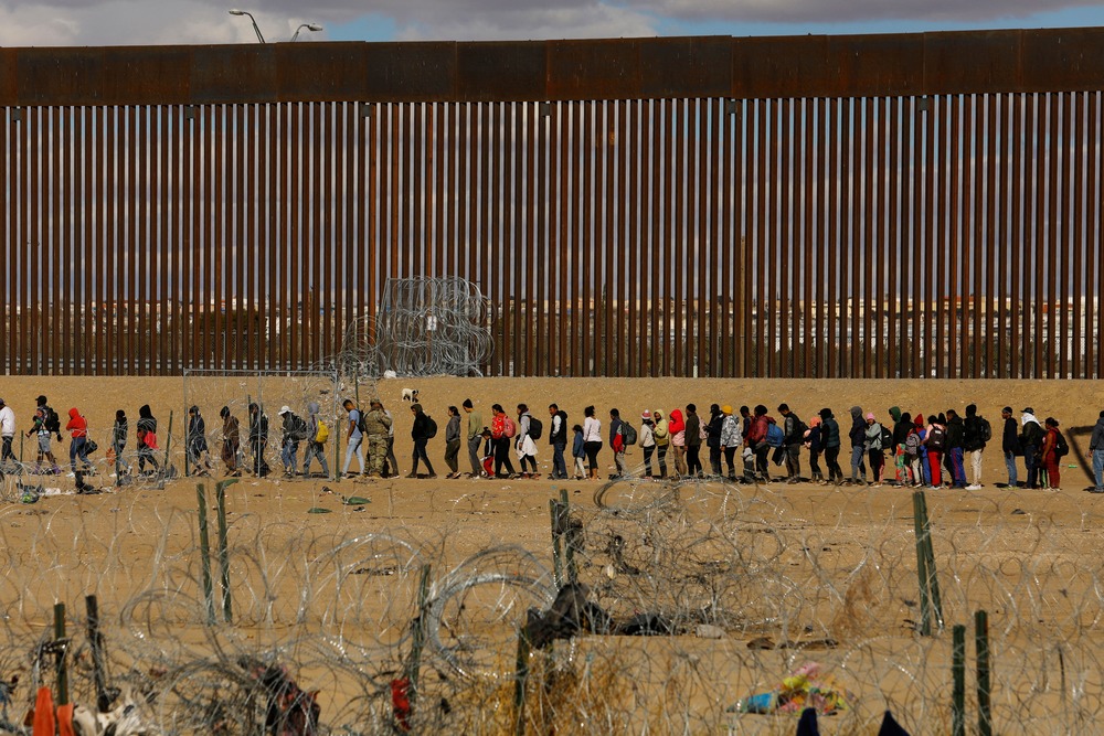 People seeking asylum lined before border wall in background.
