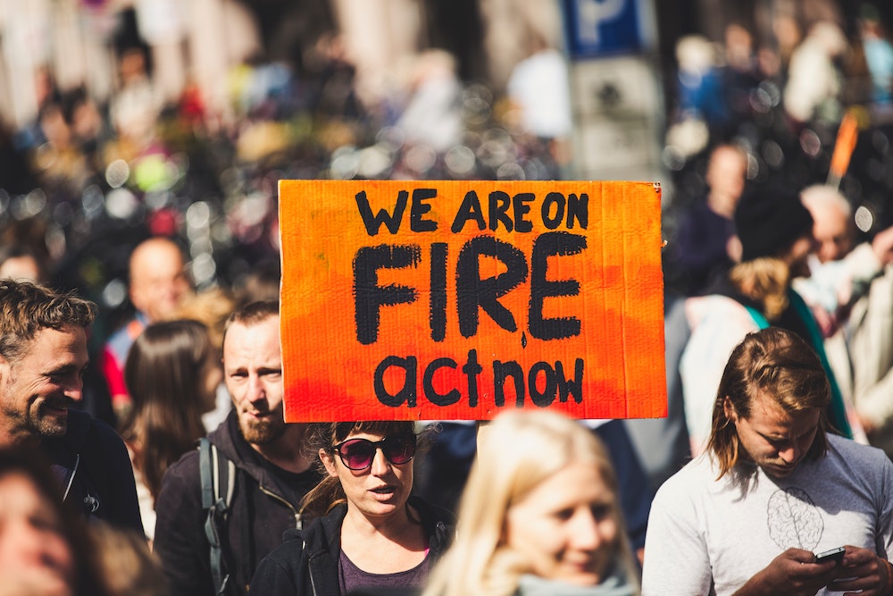 Demonstrators marching together, while a person holds a sign that reads "We are on fire, act now." (Unsplash/Markus Spiske)