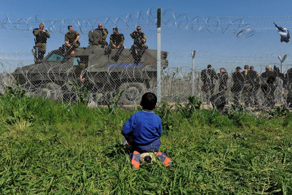 A refugee boy sits on the Greek side of the border near Idomeni as Macedonian police stand guard on their side April 12. See point three below: Pope heads to Greece. (CNS/Alexandros Avramidis)