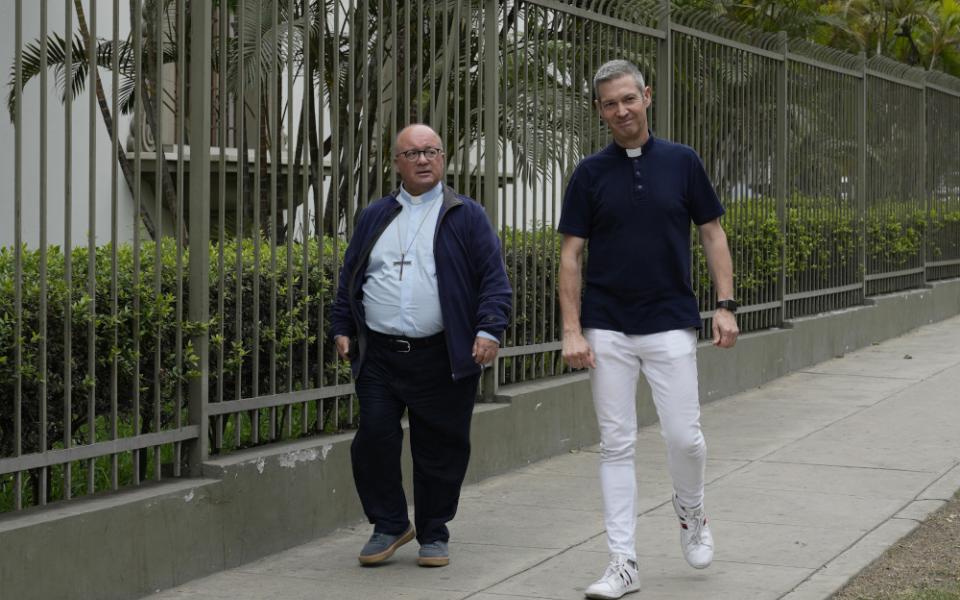 A bald man wearing a light blue shirt, clerical collar and pectoral cross walks next to a gray-haired man with a short-sleeved dark shirt with a clerical collar down a sidewalk