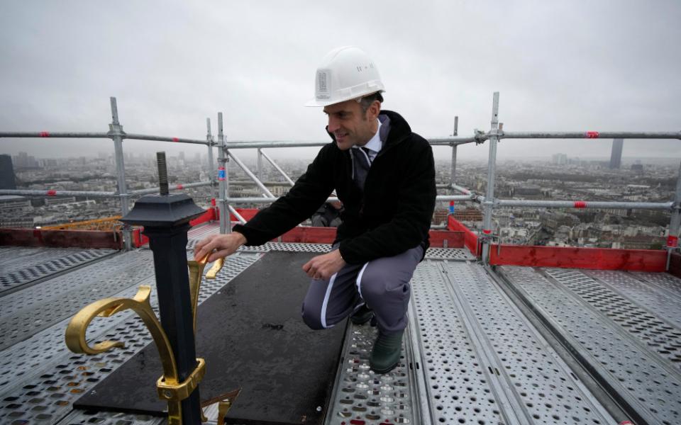 A white man wearing a white hardhat crouches on a metal platform in the sky and touches the gold top of a spire