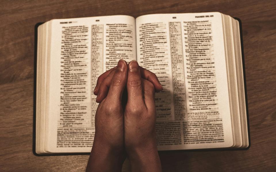 A reader holds folded hands on top of an open Bible that lays on a wooden surface. (Unsplash/Jametlene Reskp)