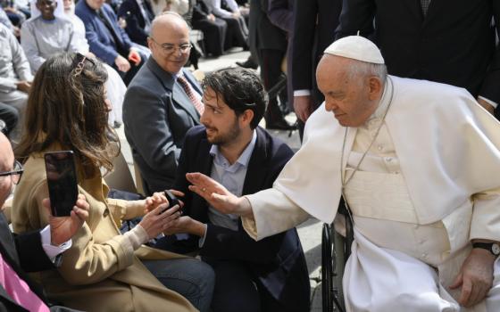 Pope Francis, seated in a wheelchair leans his hand toward a couple, where a man is kneeling with a ring box