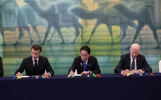 Three men in dark suits sit at a table with paper cranes on it below a painting of unidentifiable four-legged animal forms