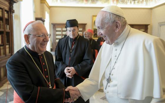 Pope Francis shakes the hand of a smiling white-haired man wearing a red zucchetto and a pectoral cross