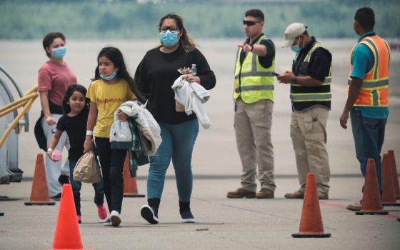 Brown people of various ages wear masks and stand on an airport tarmac next to men in safety vests