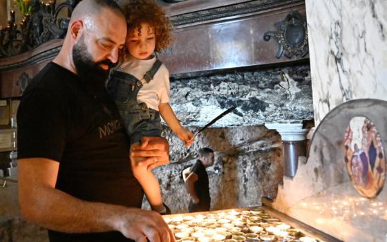 A bearded brown man holds a child in one arm and adjusts a candle with the other hand, while in a church