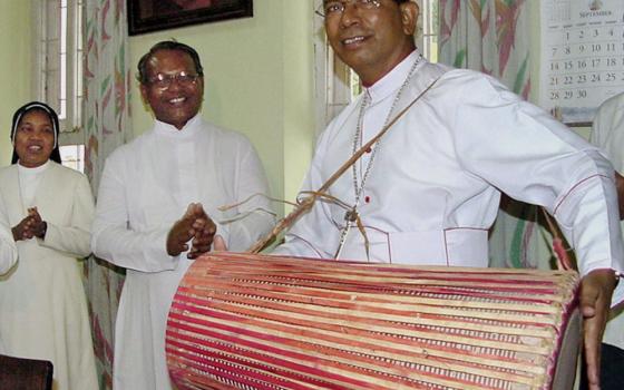 A brown man wearing white has a large drum slung over his shoulder. Another man wearing white and religious sister wearing white stand next to him.