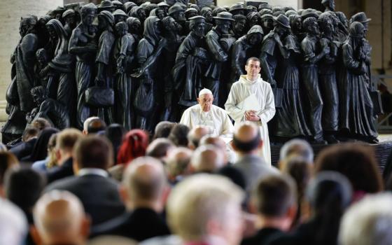 Pope Francis and a priest wearing white stand in front of metal statue of a mass of people. A crowd of people stands on the other side of them.