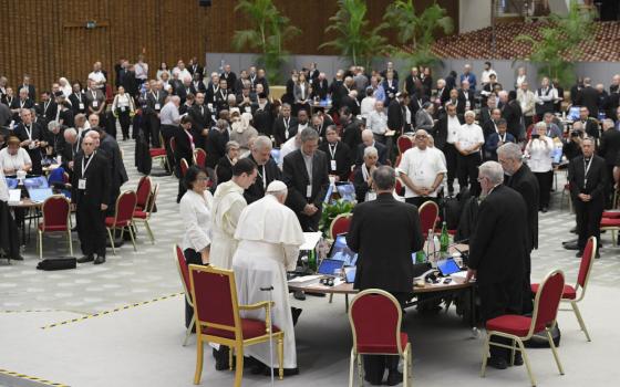 Pope Francis stands at a round table with clerics and laypeople. Other round tables are visible throughout the hall.