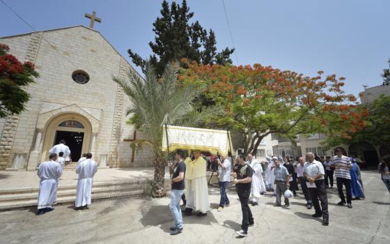 A group of people walk into a stone church. In the middle, four people hold a canopy that covers a priest holding a monstrance.