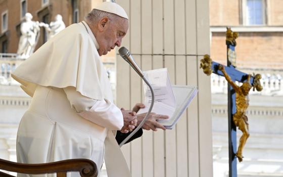Pope Francis stands, reading from notes, large crucifix hangs in background 