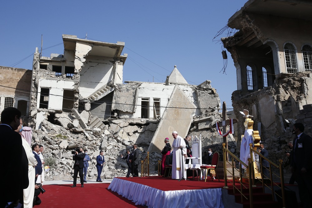 Pope Francis stands on red and white dais with rubble of bombed buildings directly behind