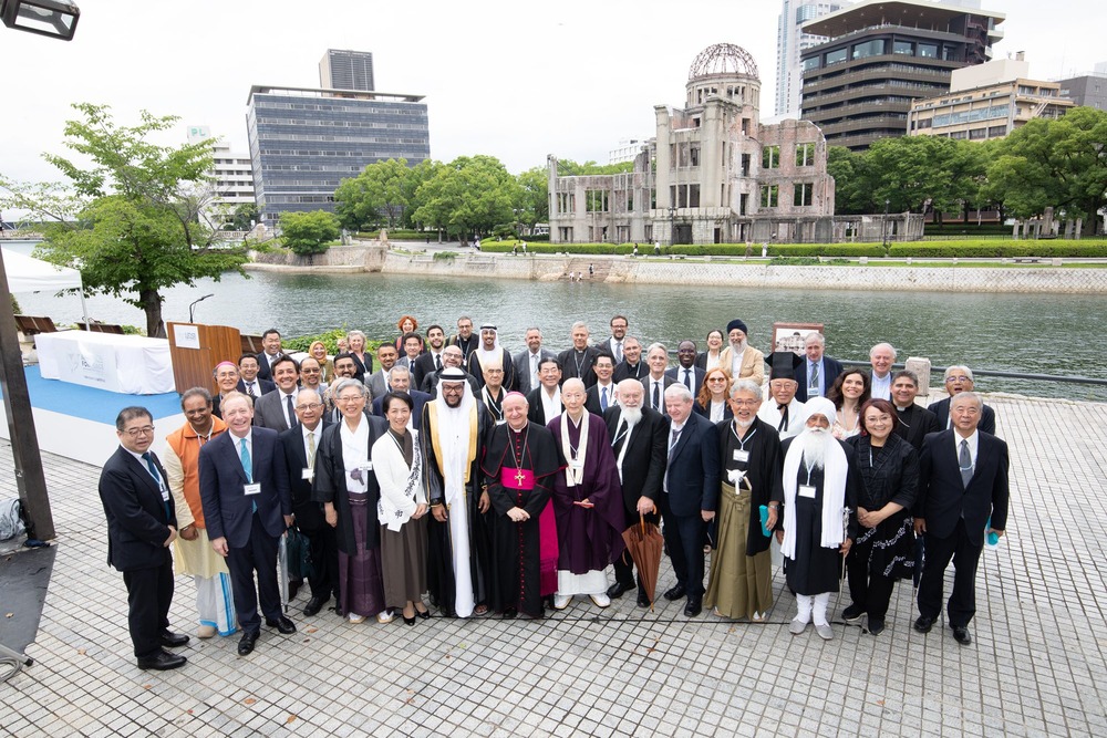 Group poses for photo before river, buildings in background. 