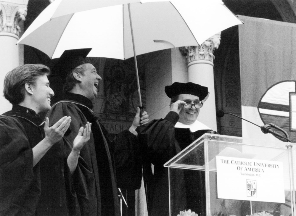 Black and white photo of the three gathered under umbrella in front of lectern. 