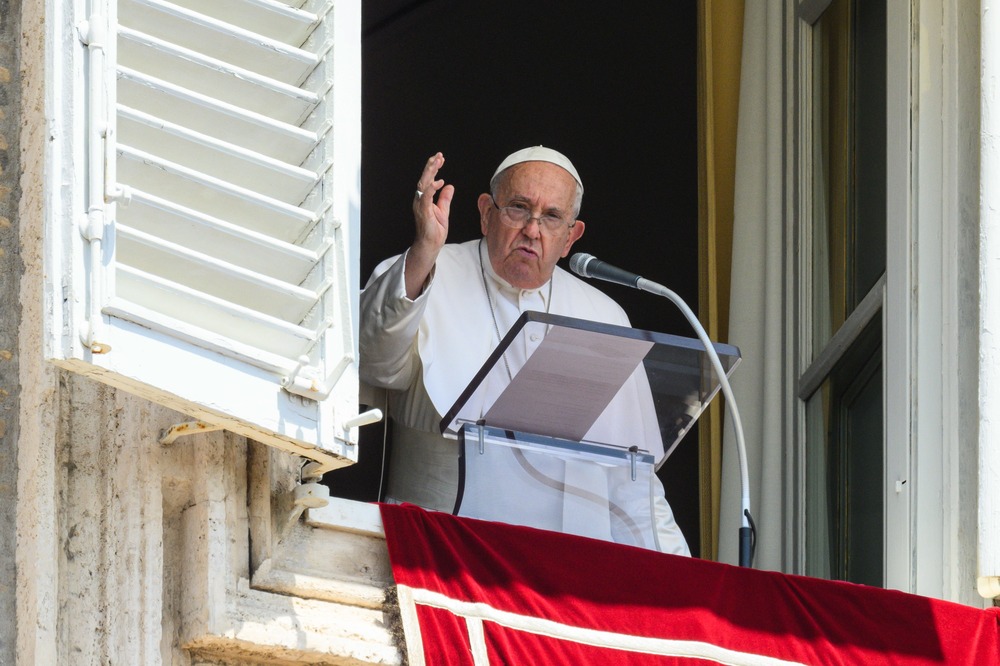 Pope Francis raises hand in blessing, standing at a lectern on the balcony in St. Peter's Square. 