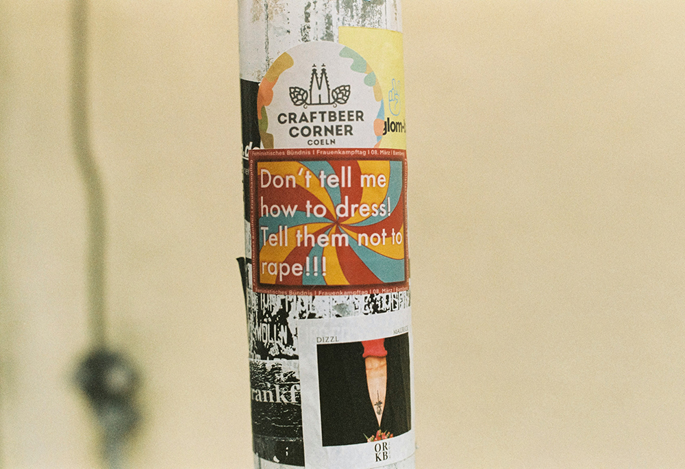 A photo shows an outside pole covered in stickers and decals. A multicolored sticker reads "Don't tell me how to dress! Tell them not to rape!!!" (Unsplash/Markus Spiske)