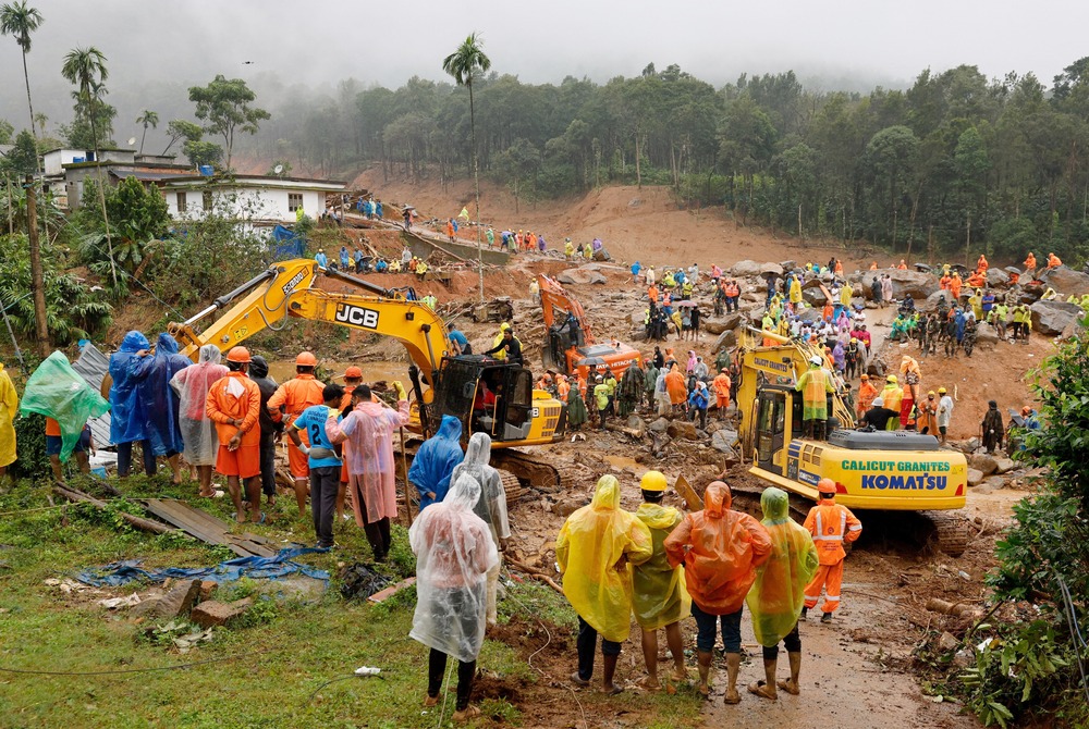 Group of people wearing rain ponchos stand around search site along with earth movers. 